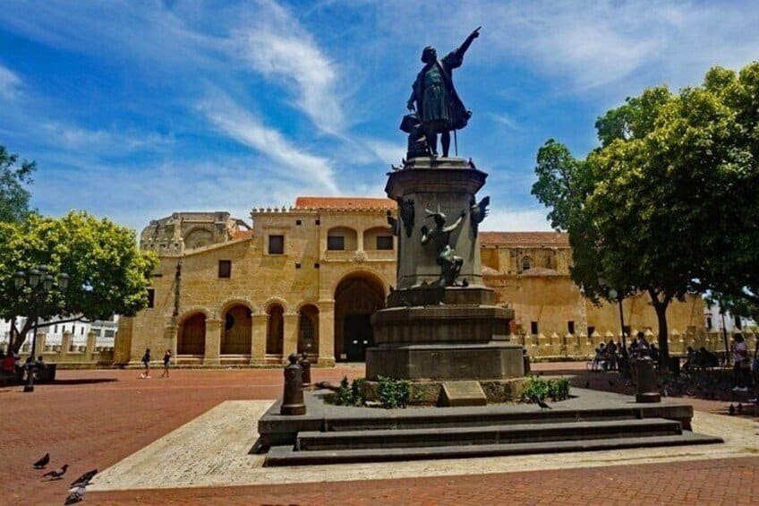 The Zona Colonial plaza in Santo Domingo, Dominican Republic, featuring a statue of Christopher Columbus standing prominently in front of the historic Catedral Primada de Am\u00e9rica, surrounded by cobblestone streets, benches, and leafy trees, with warm sunlight casting shadows across the scene.\n